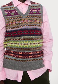 Person som bär en rosa rutiga skjorta med en flerfärgad ullstickad väst med blommiga och geometriska mönster över mörkgröna byxor.