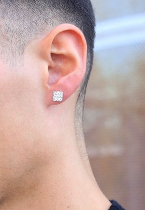 Vierkante zilveren studoorbel met een gestructureerd oppervlak, close-up van een oor. Het haar is kort geknipt rondom het oor.