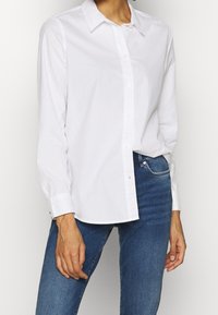 Weißes Baumwoll-Hemd mit Knöpfen, das einen klassischen Kragen und lange Ärmel hat, kombiniert mit blauen Jeans. Einfaches Design, ohne Muster oder Akzente.