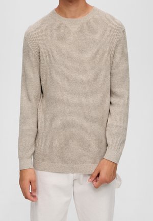 Beżowy sweter z długim rękawem o fakturowanej powierzchni, noszony przez osobę o jasnej karnacji, w połączeniu z kremowymi spodniami na jednolitym tle.