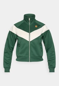 Grønn track-jakke med høy krage, hvit chevron-design, glidelås foran og ribbestrikkede ermer. Har et logo-merke på brystet.