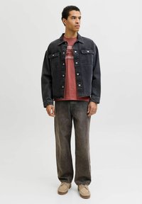 Hombre con chaqueta de mezclilla negra, jeans negros desgastados, una camiseta gráfica roja y zapatos beige, de pie contra un fondo claro y liso.