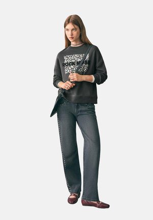 Junge Frau in einem schwarzen Sweatshirt mit weißem Text und Leopardenmuster, dunklen Jeans mit Seitennieten, burgunderfarbigen Ballerinas und einer Umhängetasche.