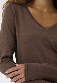 Moteris su šviesiai rudaisiais odos tonais, vilkinti rudą V formos megztinį, sudėjus rankas priekyje, demonstruojanti megztos medžiagos tekstūrą.