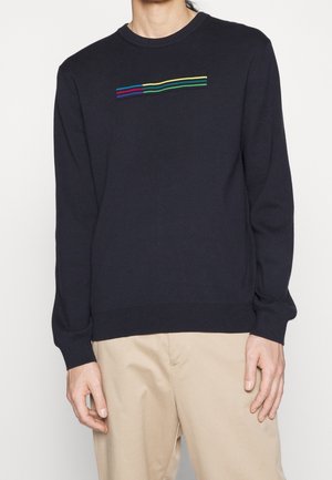 Personne portant un pull noir à manches longues avec de courtes rayures horizontales multicolores sur la poitrine et un pantalon beige.
