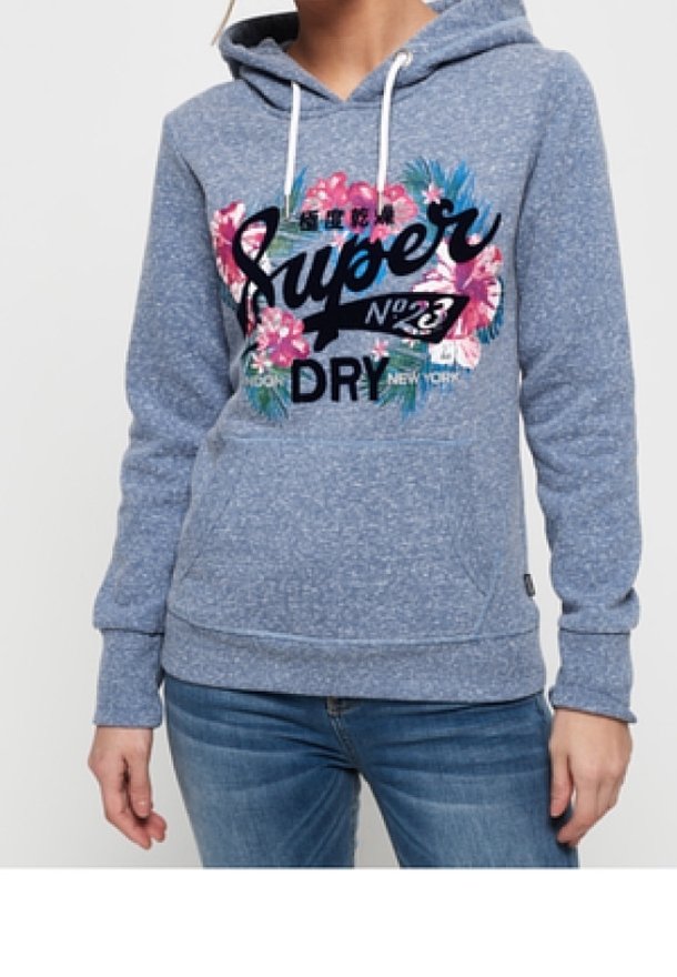 Femme portant un sweat à capuche bleu avec des cordons blancs, un motif floral et le texte "Super Dry No. 23" sur le devant, associé à un jean bleu.