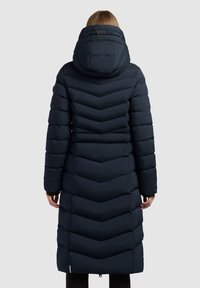 Tamsiai mėlynas prailgintas pufferis su zigzago raštu, nuimamu kapuciu ir priekiniais užtrauktukais. Ypatybės: elastingi rankogaliai ir logotipo akcentas ant rankovės.