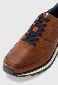 Braune Leder-Sneaker mit marineblauen Schnürsenkeln, weißer und schwarzer Sohle sowie einem strukturierten Gummikappen, schräg auf weißem Hintergrund.