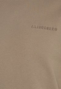 Camiseta de tela color beige con una textura suave y un sutil acanalado, con el nombre de la marca "J. Lindeberg" bordado en un hilo de contraste.