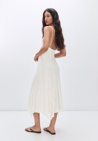 Abito midi bianco e testurizzato con corpino aderente e orlo svasato, caratterizzato da spalline sottili e una parte posteriore leggermente più lunga. Sandali completano il look.