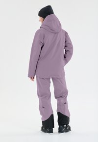 Giacca isolata viola con cappuccio e pantaloni abbinati; presenta accenti neri ai polsini e stivali; texture liscia, vestibilità moderna.