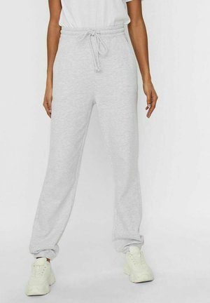 Femme portant un pantalon de jogging gris clair à cordon et des baskets blanches, debout devant un fond blanc uni.