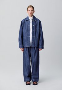 Chaqueta de mezclilla de corte relajado, de color azul claro, con costuras blancas, grandes bolsillos y cierre de botón, combinada con pantalones de pierna ancha a juego.