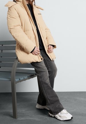 Person trägt eine beige Steppjacke, ein schwarzes Hemd, graue Jeans und weiße Turnschuhe und steht neben einer grauen Bank auf einem grauen Boden vor einer weißen Wand.