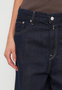 Tmavomodré rifle z denimu s hladkou textúrou, zdobené zlatým stehom, prednými vreckami a zapínaním na gombík v páse.