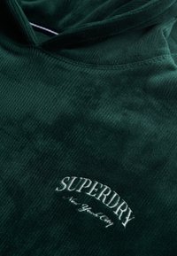 Hoodie en velours côtelé vert avec une surface texturée, arborant un logo brodé "SUPERDRY" en blanc et "New York City" en dessous.