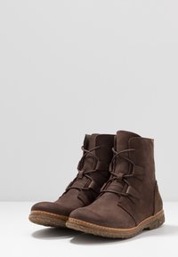 Tummanruskeat nahkaiset nilkkurit, joissa on nauhat, karkeat kumipohjat ja ommellut reunat, aseteltuna vierekkäin valkoiselle pinnalle.