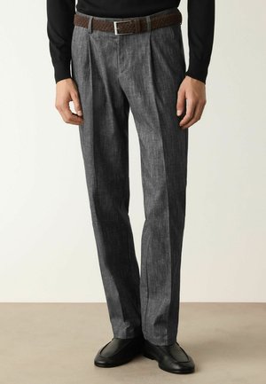 Mann trägt graue plissierte Hose mit braunem geflochtenem Gürtel und schwarzen Schuhen, steht vor einem einfarbigen hellen Hintergrund.