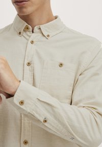 Chemise beige à boutons, en tissu texturé, avec une poche poitrine et des boutons marron. Les poignets présentent un détail de couture.