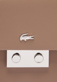 Borsa a tracolla beige con texture, caratterizzata da un logo di coccodrillo argentato e una chiusura rettangolare argentata con due accentuazioni hardware circolari.