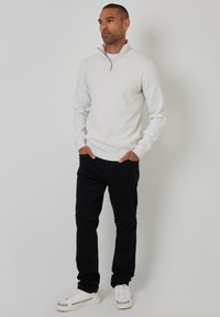 Weißer, langärmliger Reißverschluss-Pullover mit Stehkragen, kombiniert mit schwarzen Hosen und weißen Sneakers. Einfaches, strukturiertes Strickdesign.