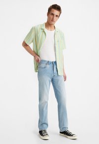 Joven con jeans de mezclilla azul claros, zapatillas negras, camiseta de tirantes blanca y una camisa de manga corta a rayas amarilla y verde, de pie contra un fondo blanco.