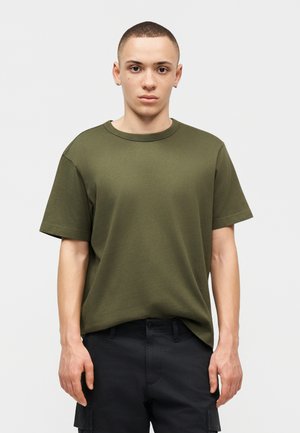 Νεαρός άνδρας με κοντά μαλλιά, ντυμένος με κοντομάνικη μπλούζα ελιάς και μαύρο παντελόνι cargo, στέκεται σε απλό φωτεινό φόντο.