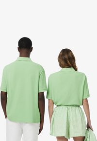 Två individer som bär matchande ljusgröna polotröjor. Tyget är slätt med en lätt lyster. Personen till höger bär också randiga shorts.