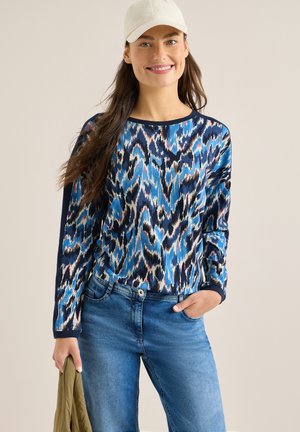 Femme souriante portant une casquette blanche, un haut à manches longues bleu à motifs, un jean bleu, tenant une veste vert olive dans une main.