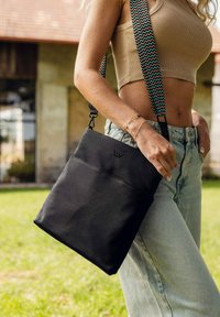 Černá látková crossbody taška s přední kapsou, která má zdobený ramenní popruh s tyrkysovým a černým zigzagovým vzorem.