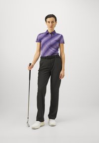 Golfantrekk med en lilla mønstrete polo skjorte og svarte bukser. Modellen holder en golfklubb og har på seg hvite golf sko.