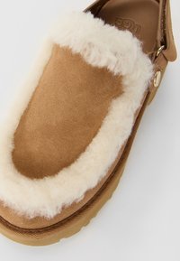 Bruine suede pantoffel met een dikke crème-kleurige wolvoering en zool, voorzien van een bandje met een gouden knopelement.