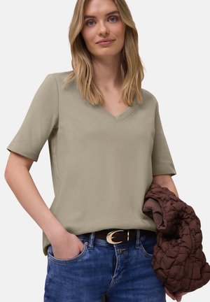 Mujer con cabello rubio hasta los hombros, vestida con una camiseta de cuello en V beige y jeans azules, sosteniendo una chaqueta acolchada marrón en una mano.
