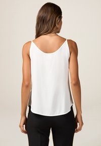 Femme aux longs cheveux bruns portant un haut blanc sans manches et un pantalon noir, vue de dos sur un fond uni.