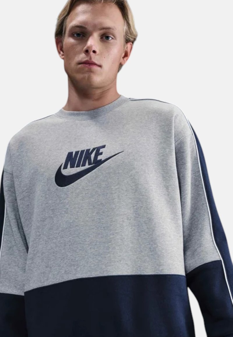 Giovane uomo che indossa una felpa Nike grigia e blu navy con il logo al centro del petto, in piedi contro uno sfondo chiaro e neutro.