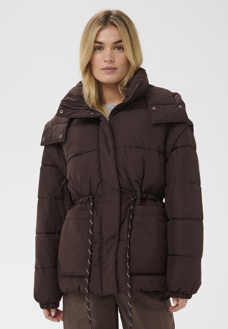 Femme portant une veste matelassée brun foncé avec col montant, poches avant et taille ajustable par cordon, sur un fond uni.