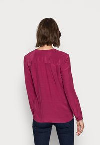 Chemise à manches longues en satin rose, avec un col rond, des fronces à l'arrière et des poignets boutonnés. Associée à un jean foncé.