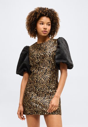 Žena s kovrčavom kosom u haljini s leopard uzorkom i velikim crnim napuhanih rukava, stoji ispred jednobojne svijetle pozadine.