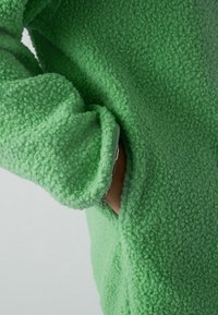Pullover in pile verde con superficie tessuta e soffice. Presenta una tasca laterale e polsini a costine per un comfort aggiuntivo.