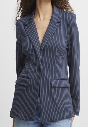 Frau trägt einen taillierten dunkelblauen Blazer mit weißen Nadelstreifen und Pattentaschen, kombiniert mit hellblauen Jeans.