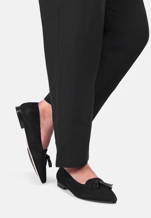 Person iført sorte loafers med spids tå og kvaster samt sorte bukser, med den ene fod løftet let bag den anden.