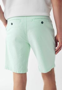 Pantaloni scurți verde mentă cu buzunare la spate și nasturi negri. Materialul pare neted, cu o ușoară strălucire și tivuri rulate la manșete.