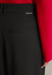 Pantalones negro a rayas con un bolsillo y una etiqueta que dice "Marc O'Polo DENIM". La tela texturizada contrasta con un suéter rojo.