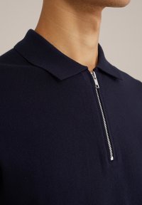Polo de punto azul marino con cuello con cremallera. La tela tiene una textura suave y presenta un detalle de cremallera plateada en el cuello.