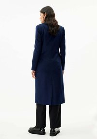 Lång marinblå kappa med en figurvänlig silhuett, skräddarsydd design, långa ärmar, mitt bak-ventilation och slät textur, tillsammans med svarta byxor och skor.