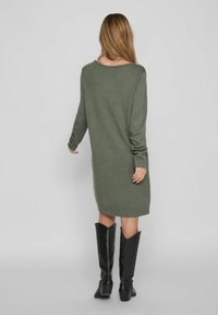 Långärmad, oversized tröjklänning i olivgrön, ribbade ärmslut och nederkant, med en båtkrage och mjuk stickad textur. Bärs med svarta knähöga stövlar.
