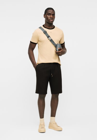 Άνδρας που στέκεται φορώντας μπεζ κοντομάνικη μπλούζα με μαύρη λεπτομέρεια, μαύρο σορτς, μπεζ αθλητικά παπούτσια και γκρι τσάντα ώμου, μπροστά σε απλό λευκό φόντο.