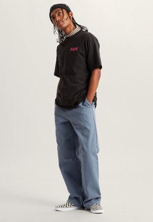 Sort bomulds-T-shirt med broderet tekst, kombineret med lyseblå, løstsiddende bukser. Modellen bærer sorte og hvide ternede slip-on sko.