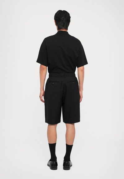 Chemise noire à manches courtes associée à un short noir, présentant une taille élastique et deux poches arrière, portée avec des chaussettes et des chaussures noires.