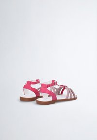 Vaaleanpunaiset sandaalit, joissa on yhdistelmä sileitä ja teksturoituja hihnoja, säädettävä nilkkahakas ja litteä ruskea pohja.